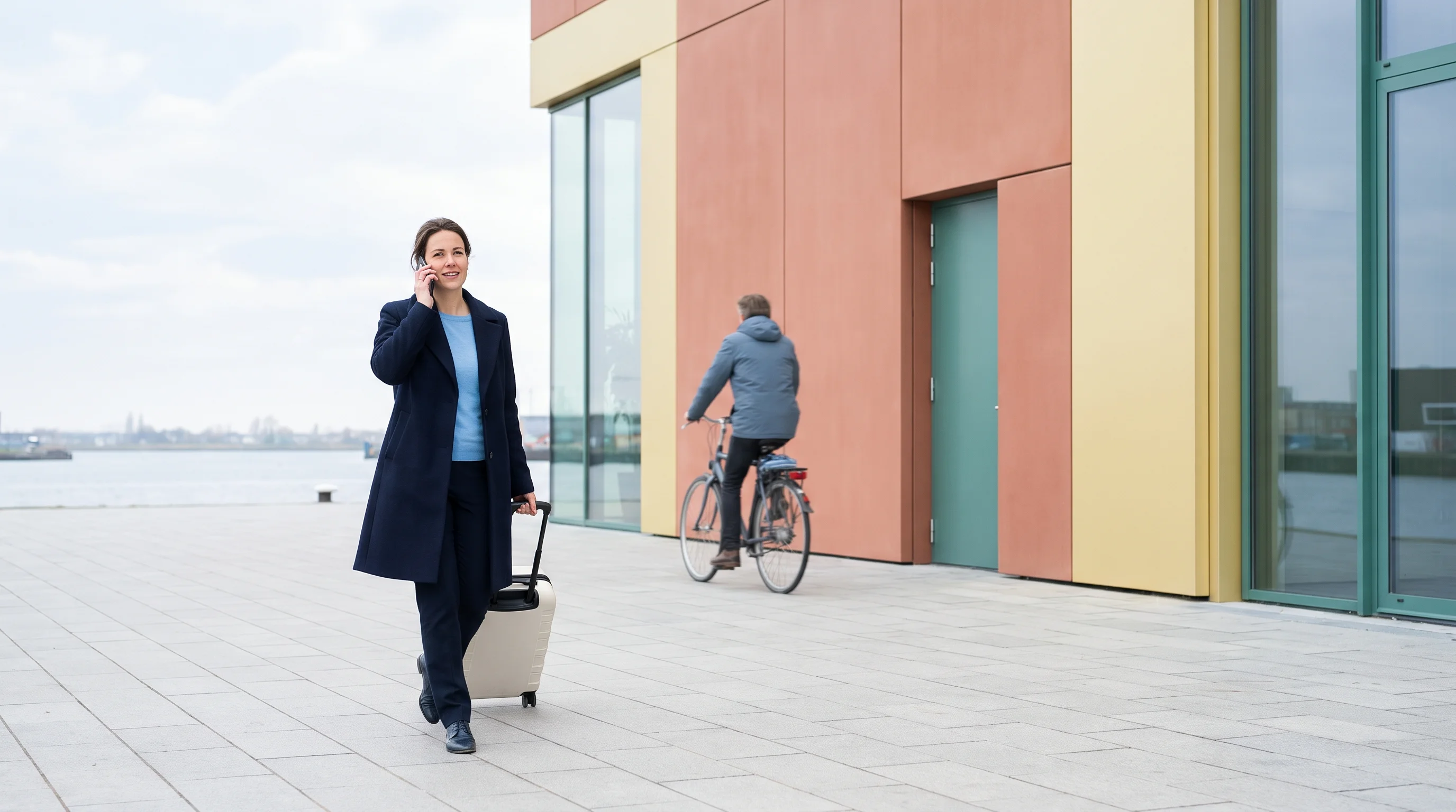 Een vrouw belt terwijl ze met haar koffer langs een modern gebouw aan het IJ loopt