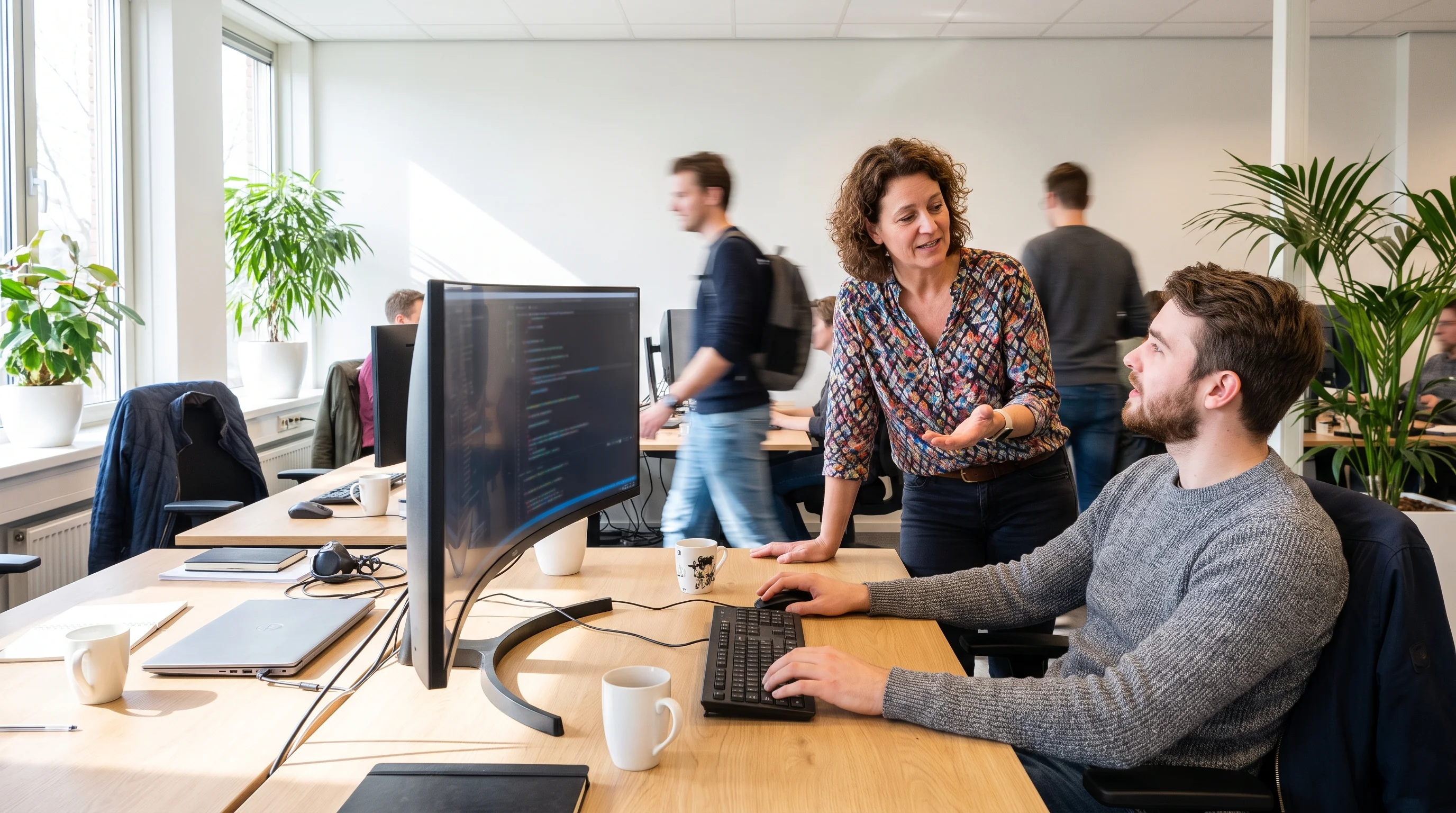 An experienced developer stands next to a young colleague at his desk, talking him through the code on his screen