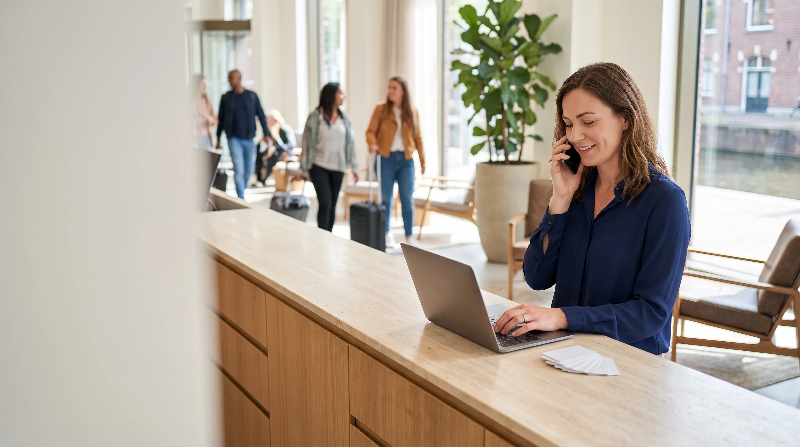 A receptionist in a sunny hotel lobby calls the helpdesk while working on her laptop; guests with suitcases walk past behind her