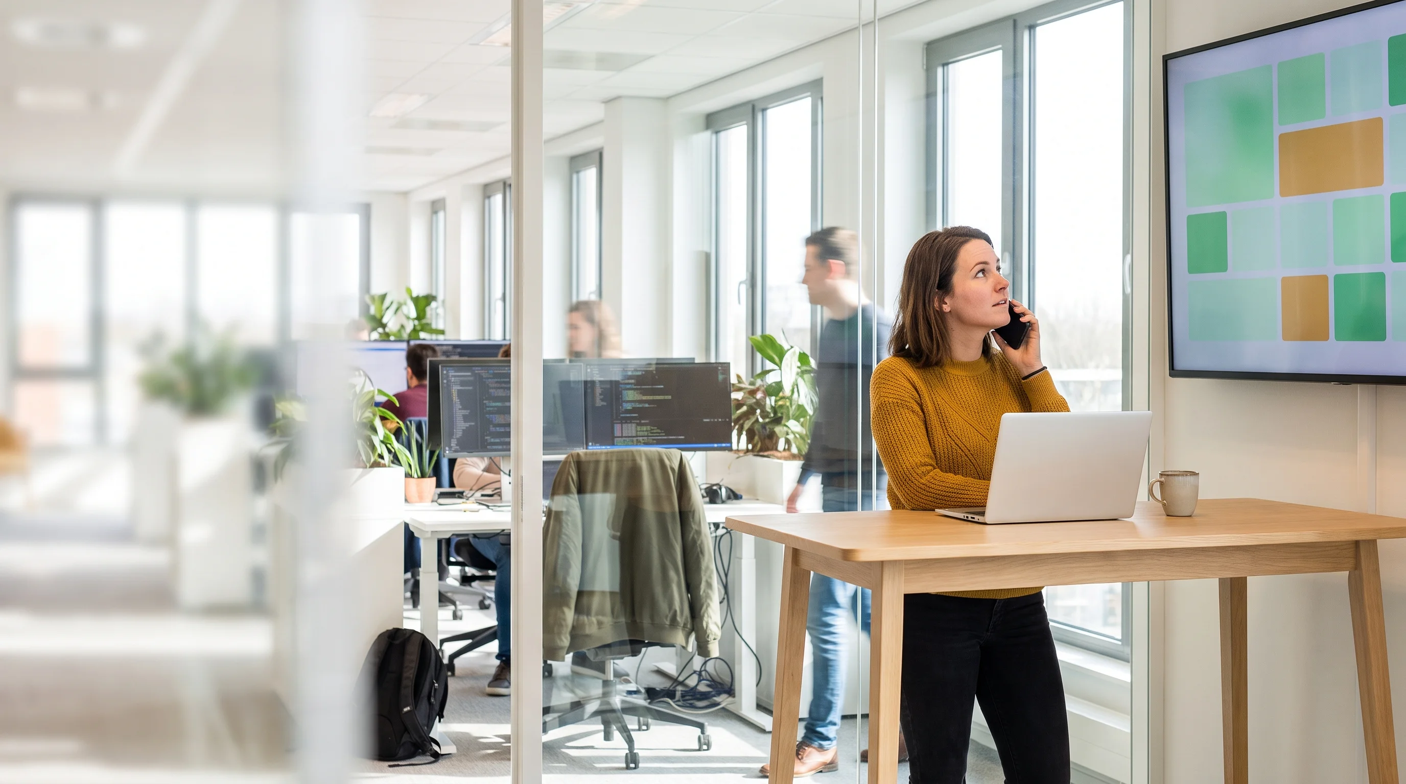 A DevOps engineer on a call at a standing desk with her laptop, looking at a status dashboard on the wall