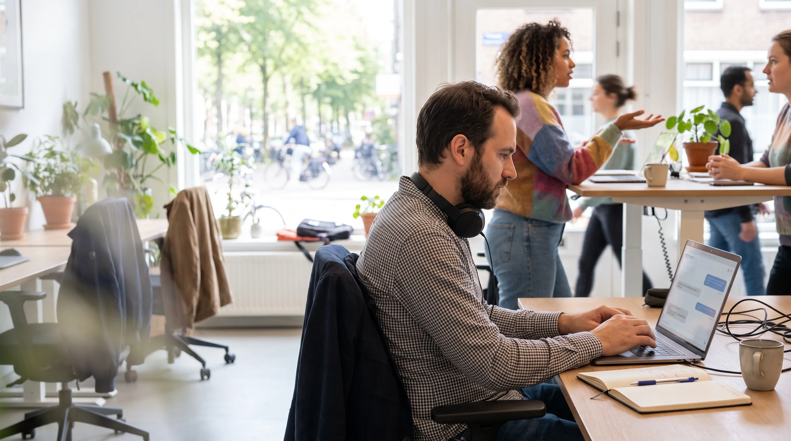 A developer with headphones round his neck works intently on his laptop, with code and a chat window, in a sunny workspace
